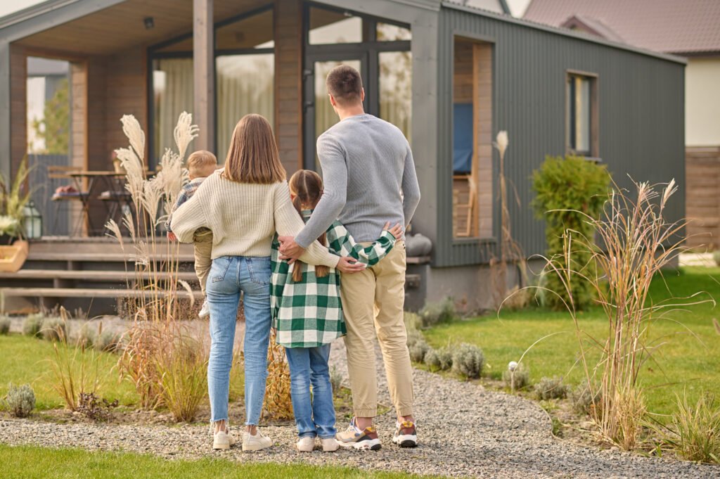 Happy family. View from back of man woman with child and girl standing together hugging and admiring their cozy home on fine autumn day